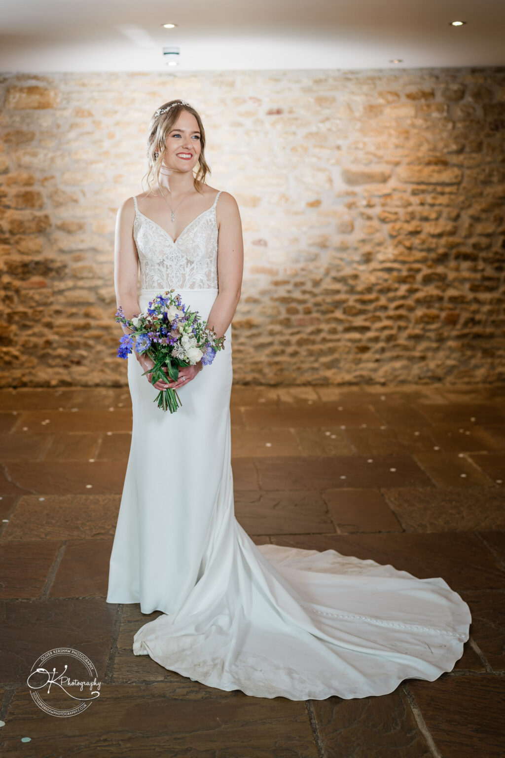 A portrait of the bride in her wedding dress holding a bouquet, standing against a stone wall at Dodford Manor.