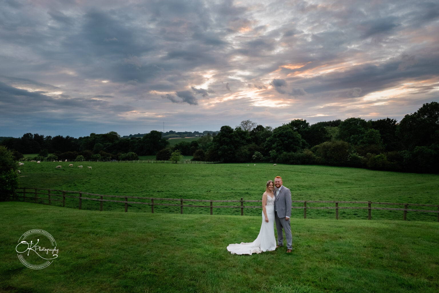 Couple in wedding attire posing on a grassy field with a fenced pasture, trees, and a cloudy sky in the background.
