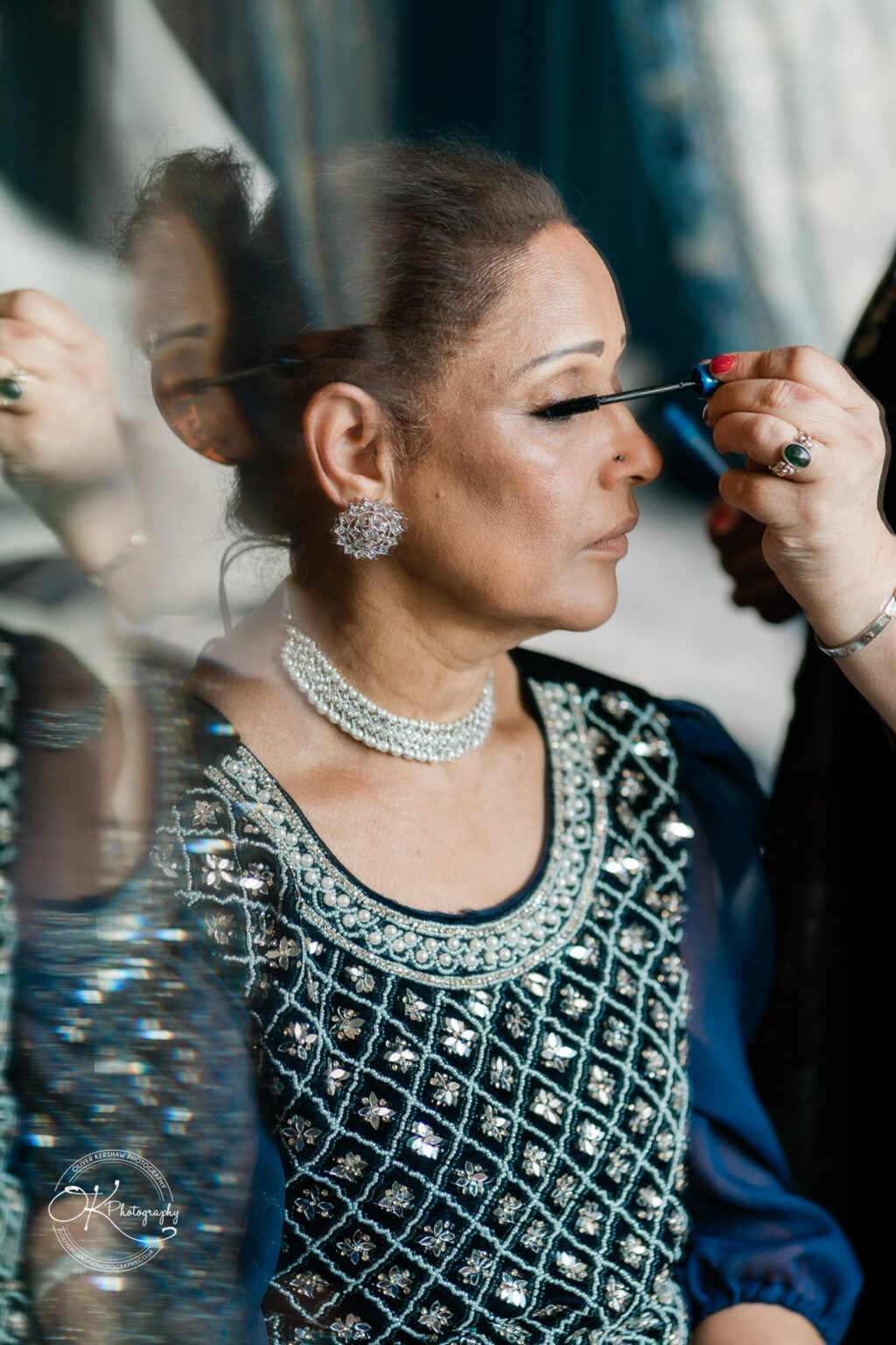 Elegant woman in traditional attire having her makeup done, focusing on mascara application, with a soft reflection effect.