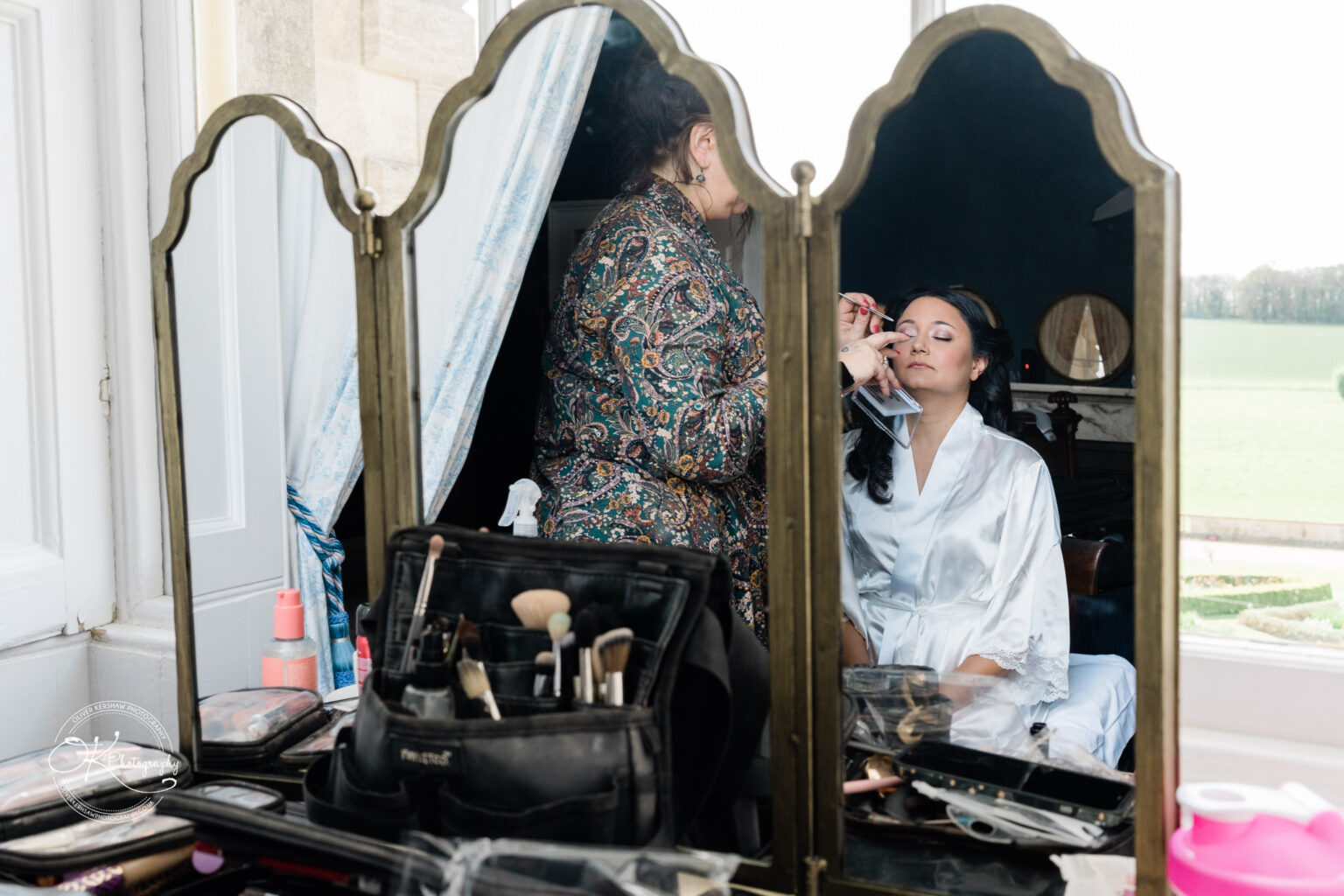Bride having makeup applied while seated in front of a mirror with makeup tools visible.