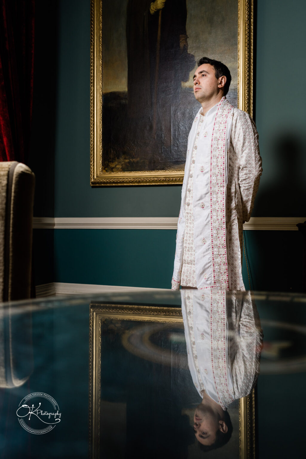 Groom in traditional attire standing reflectively in front of a portrait painting, with a reflection below.