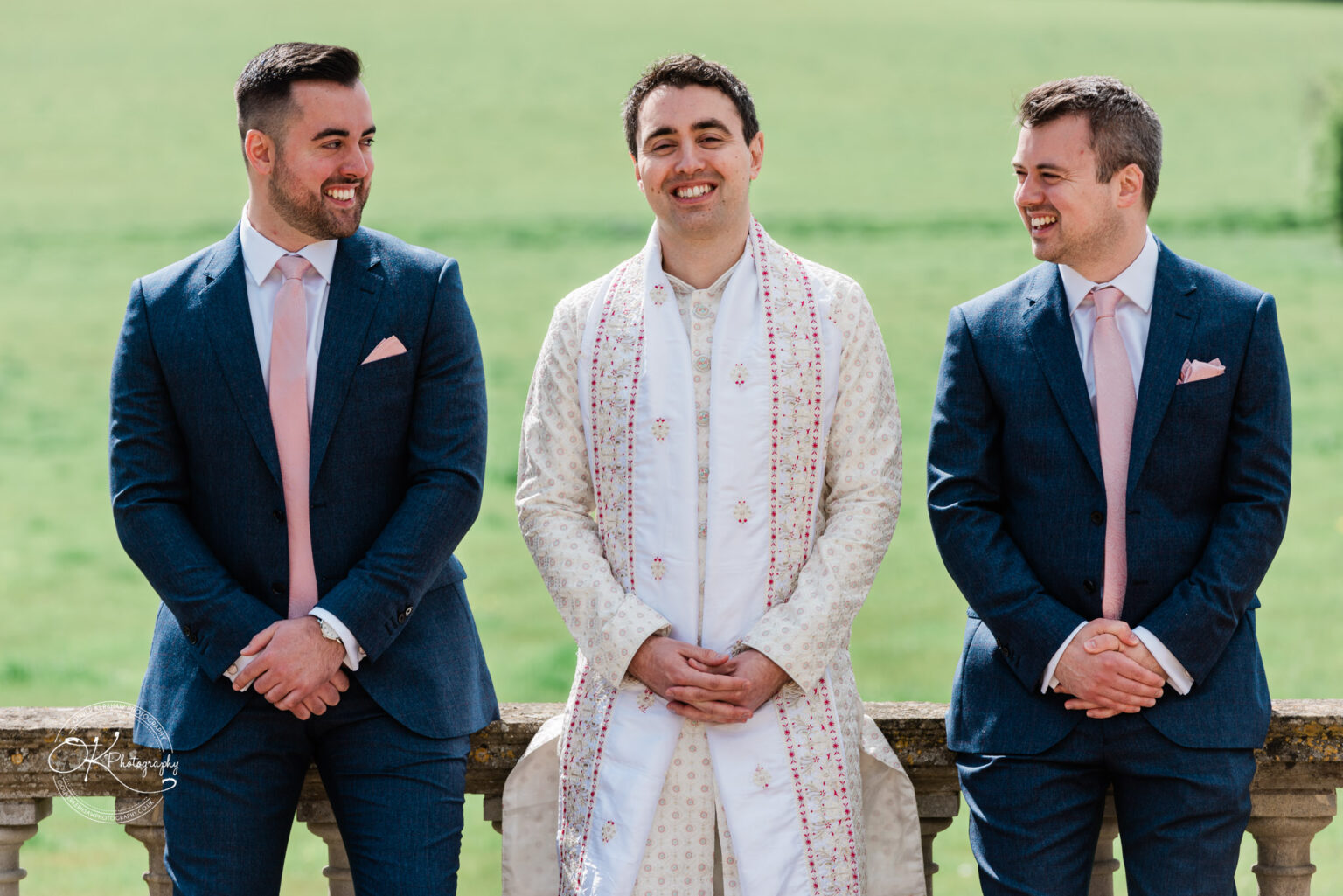 Groom with two groomsmen, dressed in suits and traditional attire, smiling outdoors.