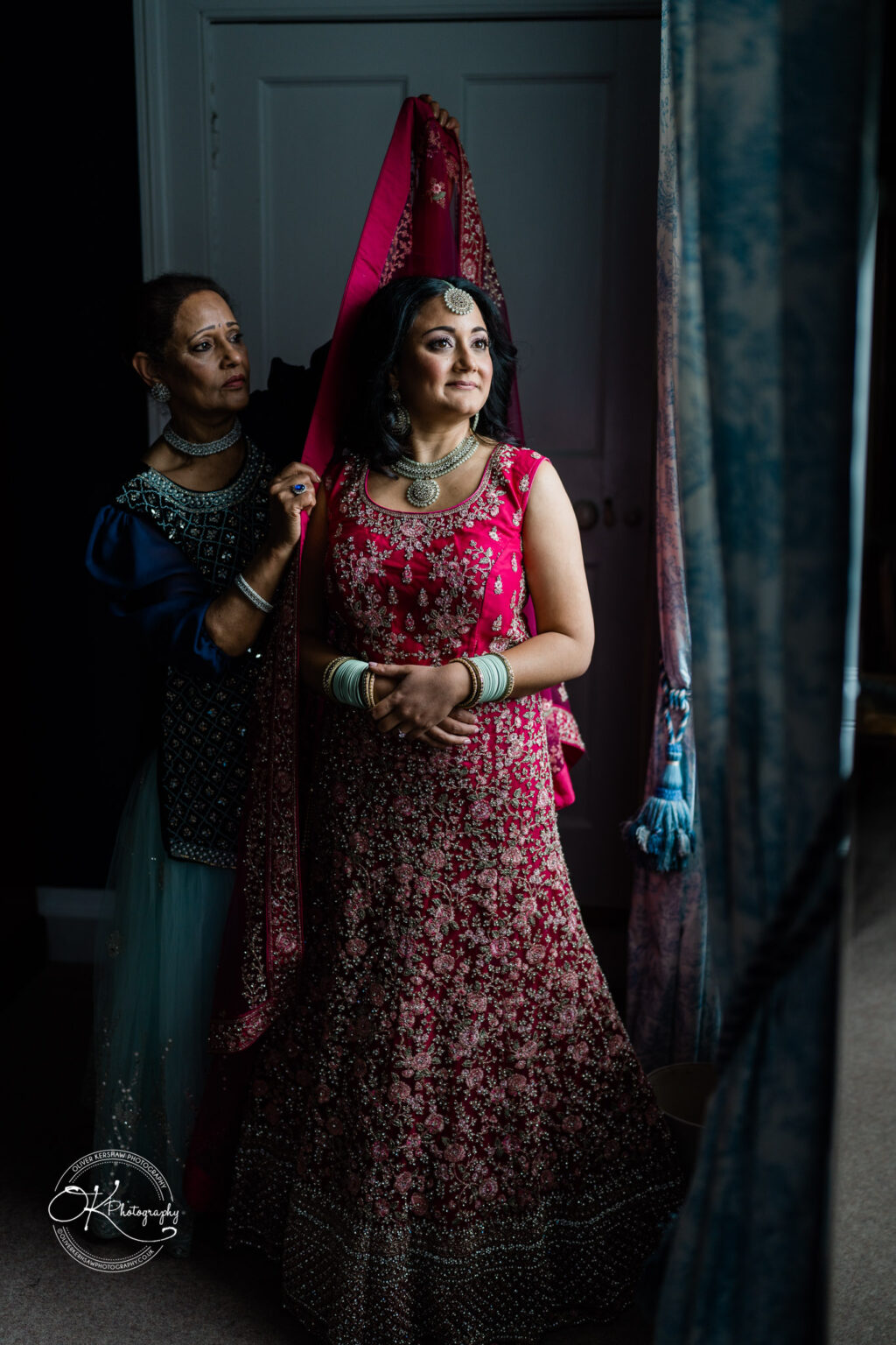 Bride standing with an older woman helping to adjust her wedding attire, captured in a softly lit room.