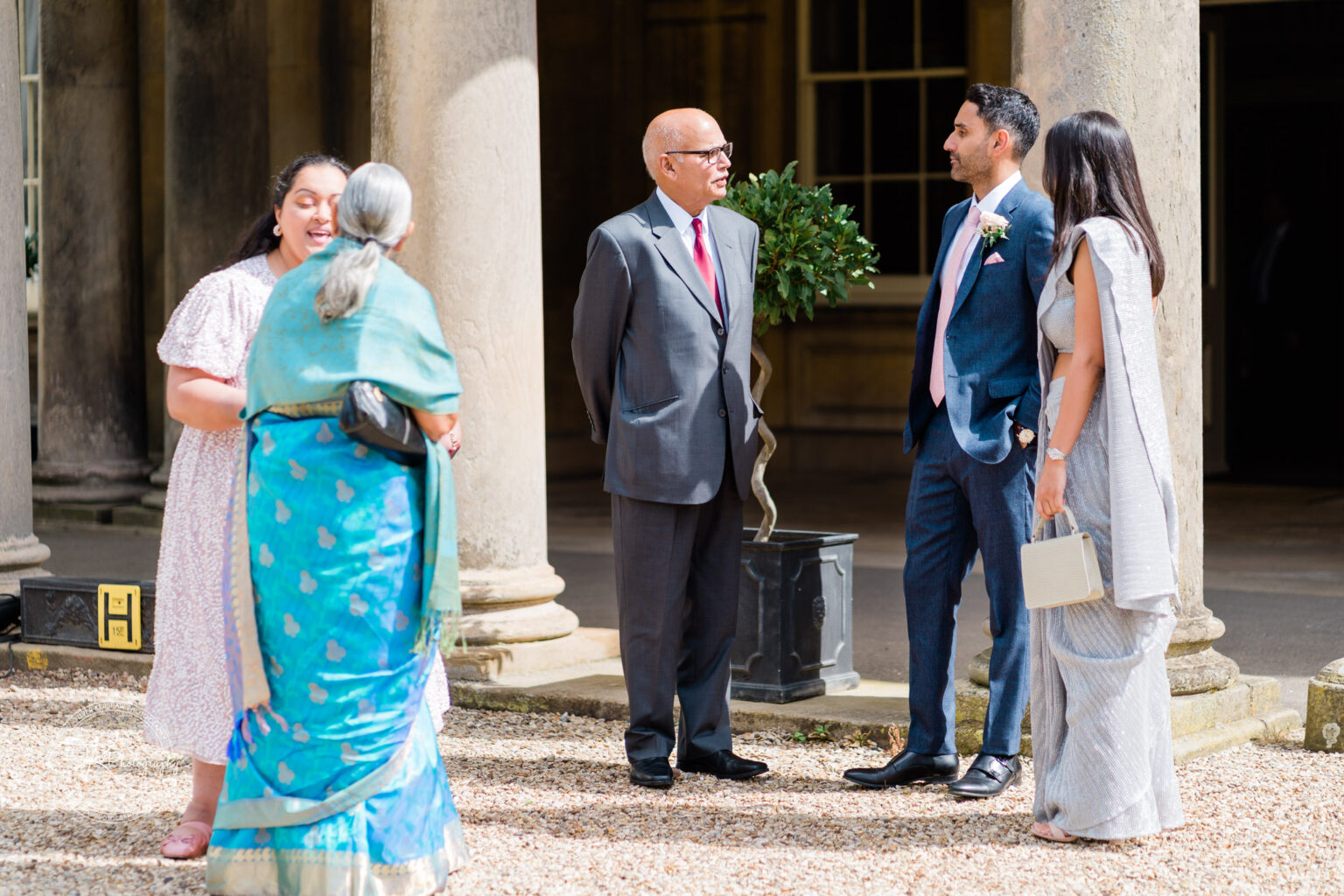 Guests in formal attire engaging in conversation outside a grand venue, with columns in the background.
