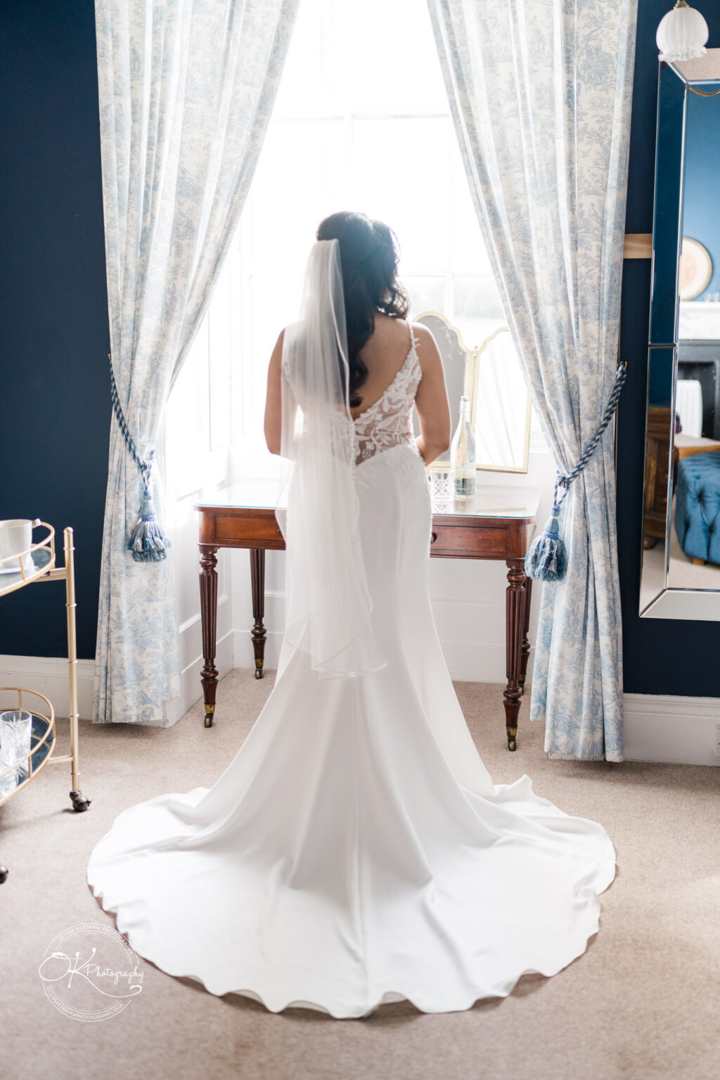 Bride in white gown facing a window, looking out with sunlight streaming in.