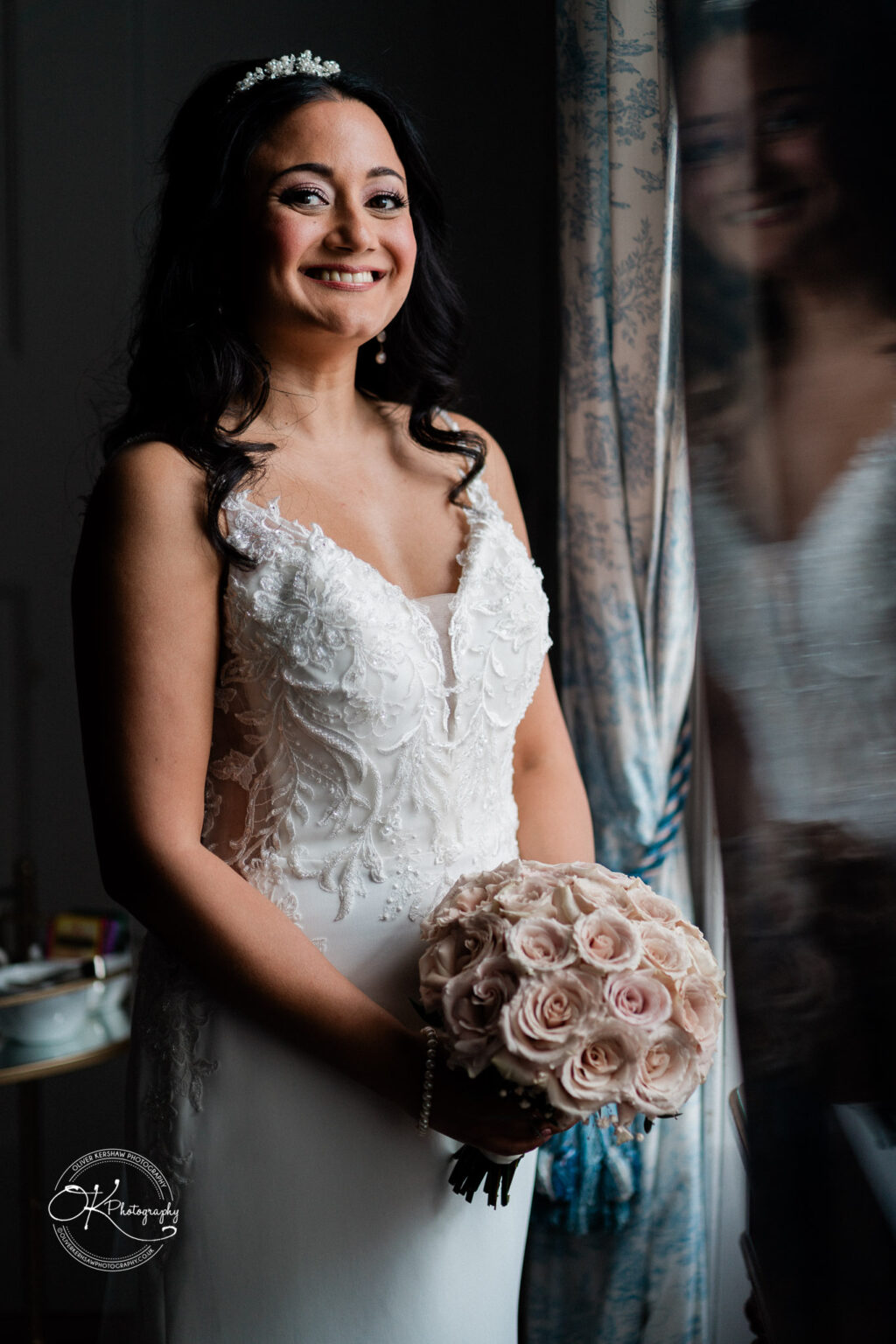 Bride smiling while holding her bouquet, standing near a window.