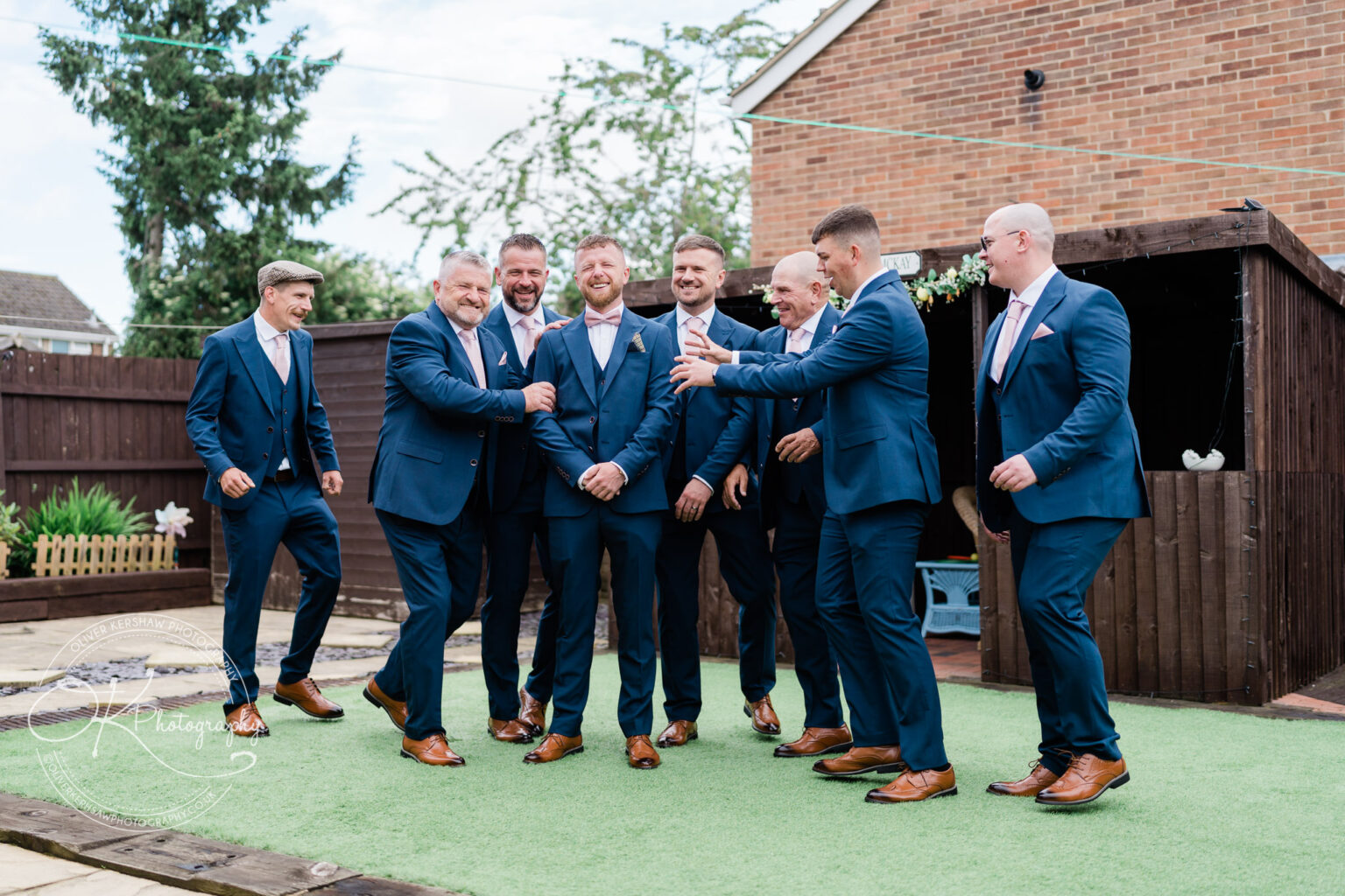 A group of men in matching navy blue suits and pink ties laughing and enjoying themselves in a backyard.