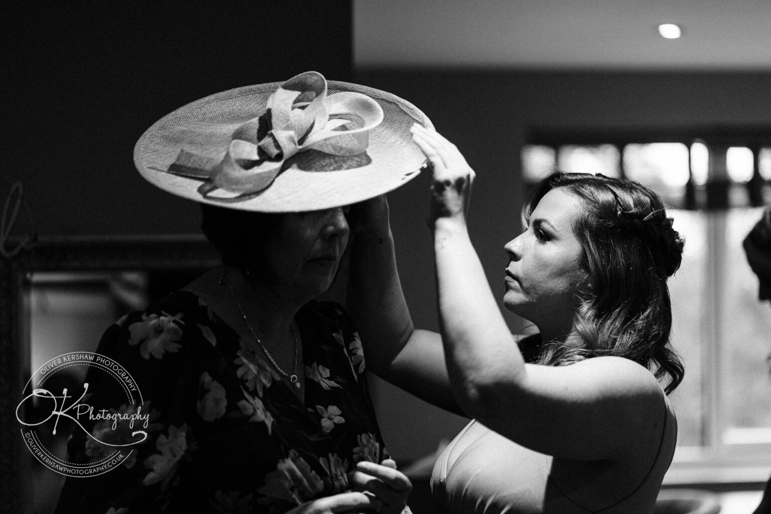 A woman helps another woman adjust a large hat with an ornate bow in a dimly lit room.