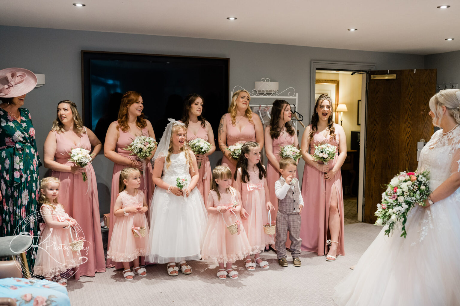 Bridesmaids and flower girls in pink dresses and holding bouquets, along with a bride in a white gown, stand together smiling and chatting in a room.