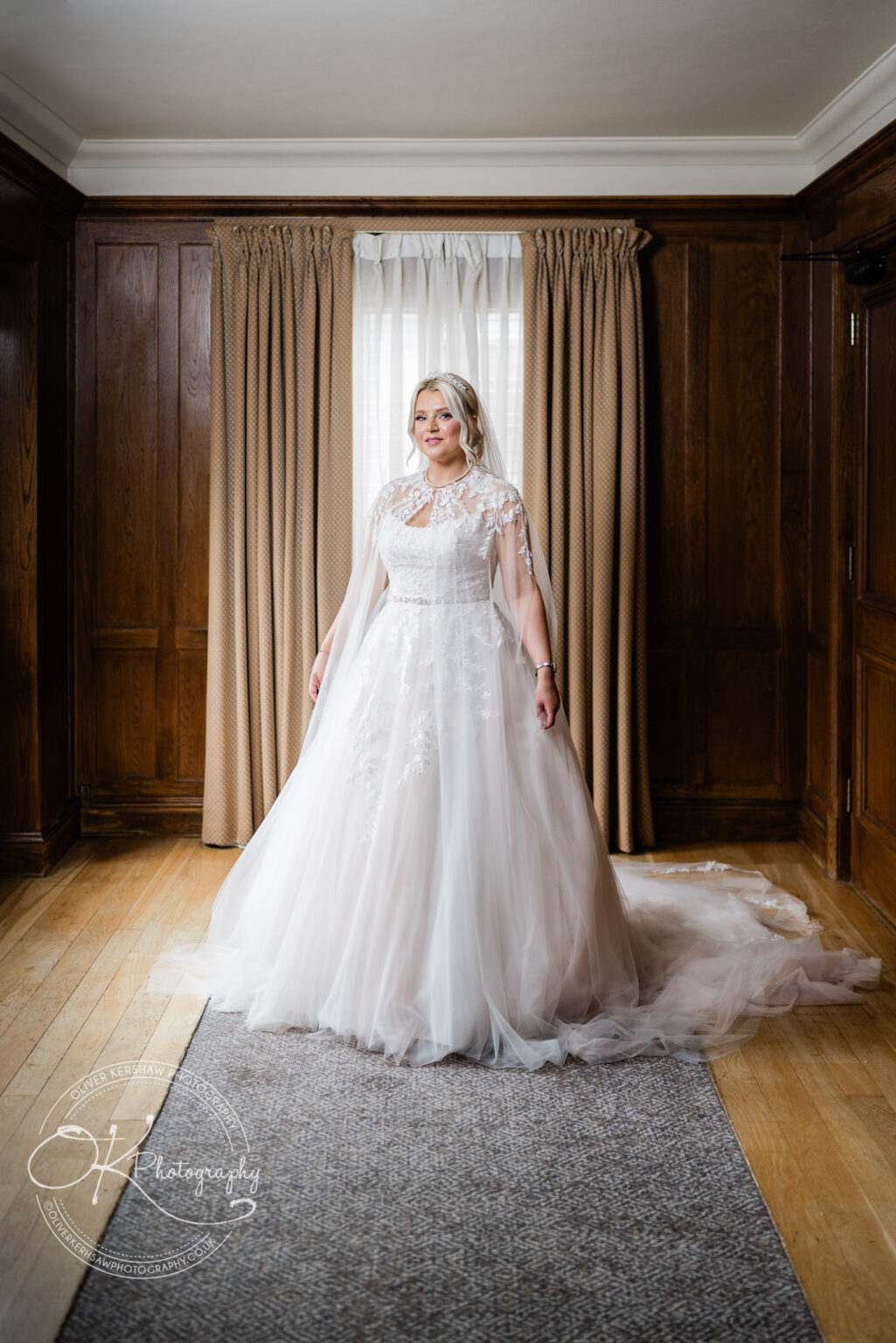 Bride in a white lace wedding dress standing in front of window curtains within a wood-panelled room.