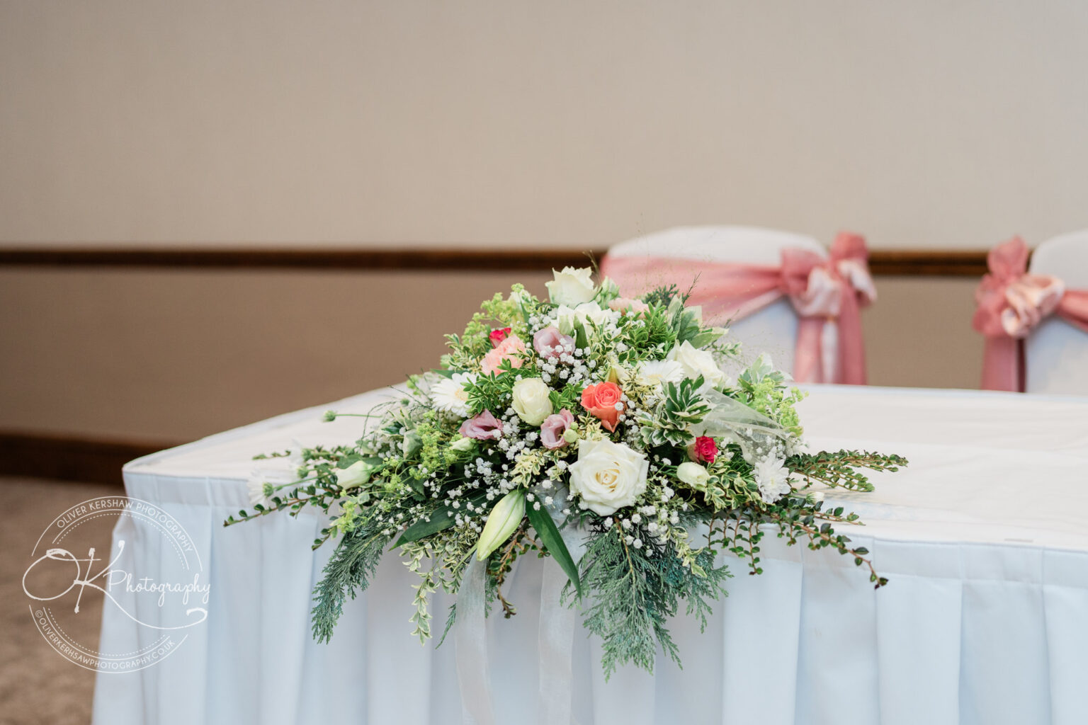 Floral arrangement with white, pink, and red flowers on a white table, with chairs adorned with pink bows in the background.
