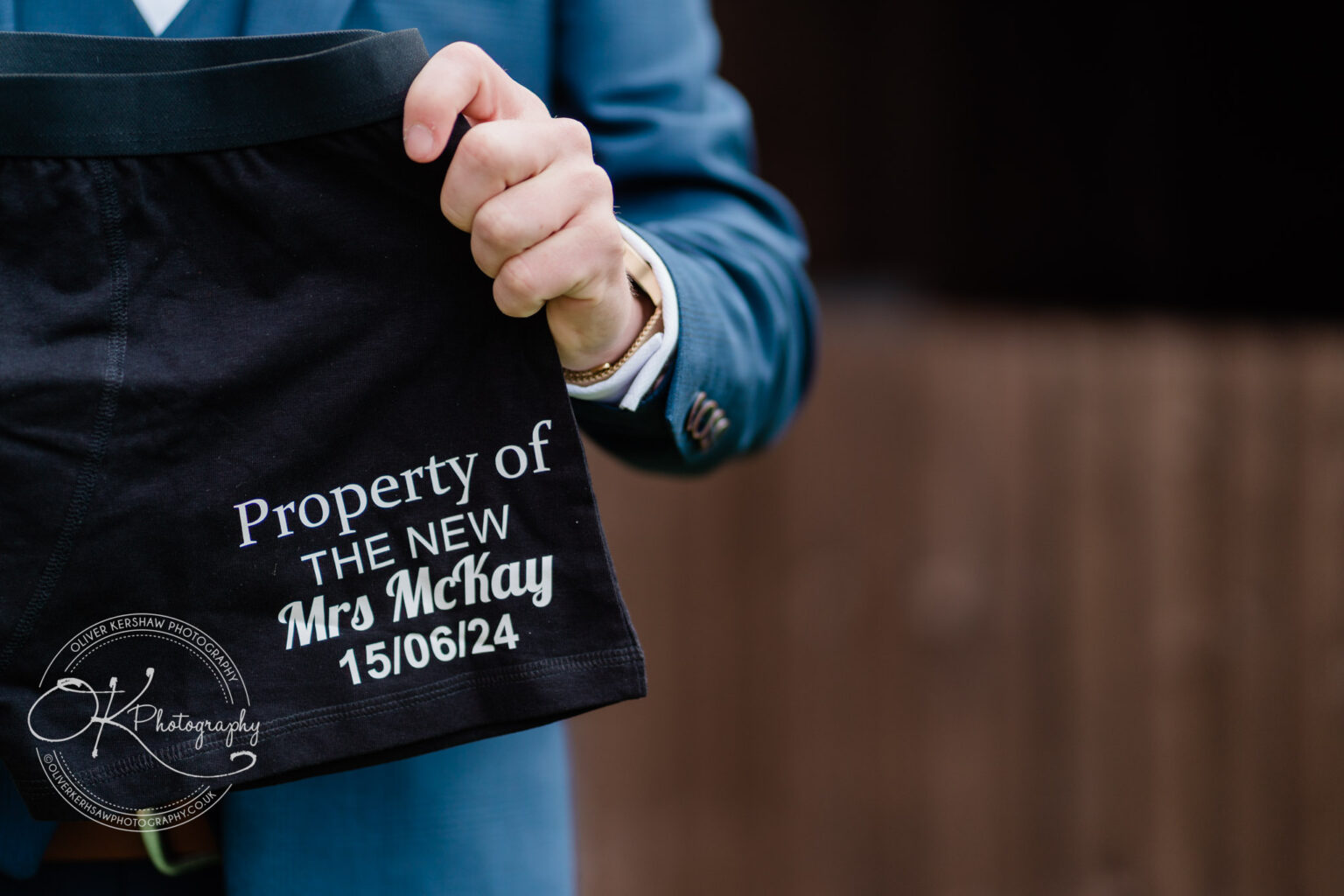 A person in a blue suit holds up a pair of black underwear with the text "Property of THE NEW Mrs McKay 15/06/24" printed on them.