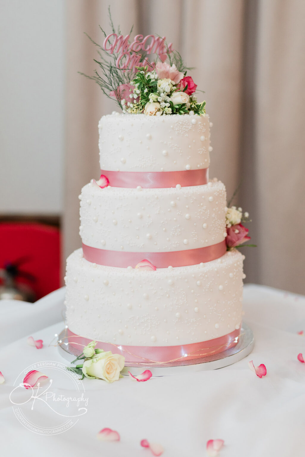 Three-tiered white wedding cake with pink ribbons and pearl decorations, topped with flowers and the text "Mr & Mrs".