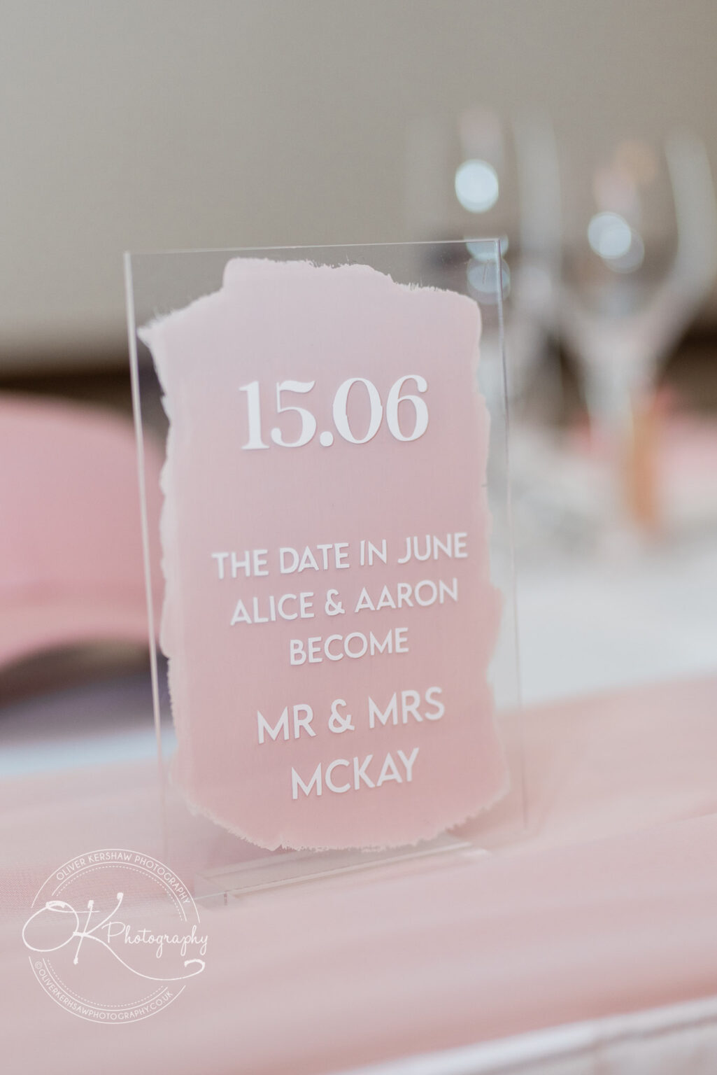 A wedding table sign with a pink background, displaying the date "15.06" and the message "The date in June Alice & Aaron become Mr & Mrs McKay."