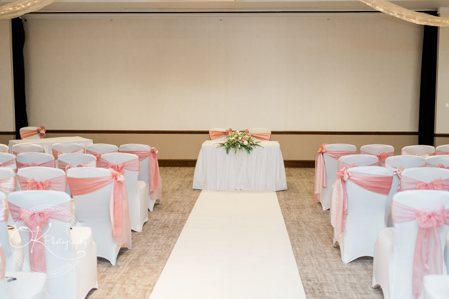 A wedding ceremony setup with a white aisle, white chair covers adorned with pink sashes, and a decorated table at the front.