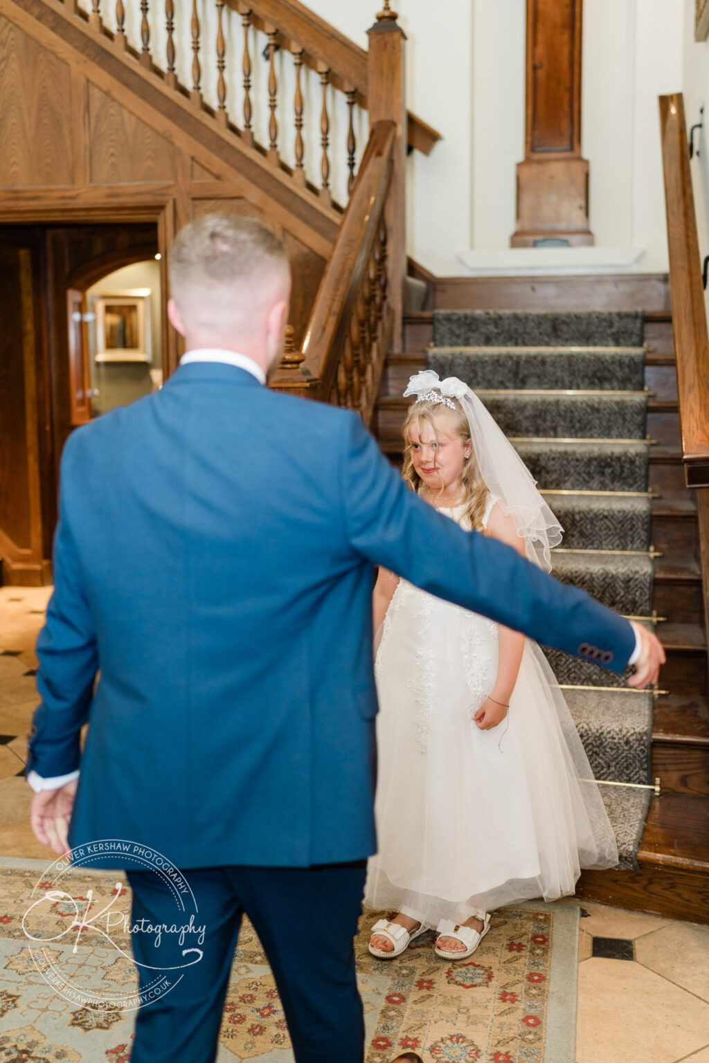 A man in a suit with arms outstretched stands in front of a young girl in a white dress and veil on a staircase.