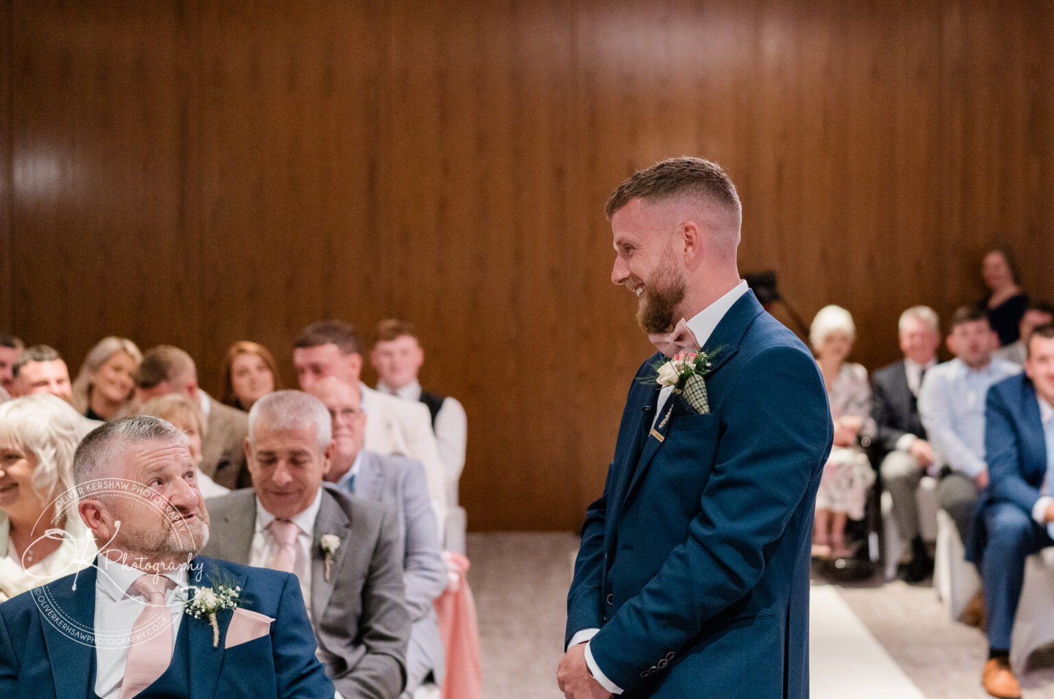 A man in a suit smiles while standing in front of a seated crowd at a wedding ceremony.