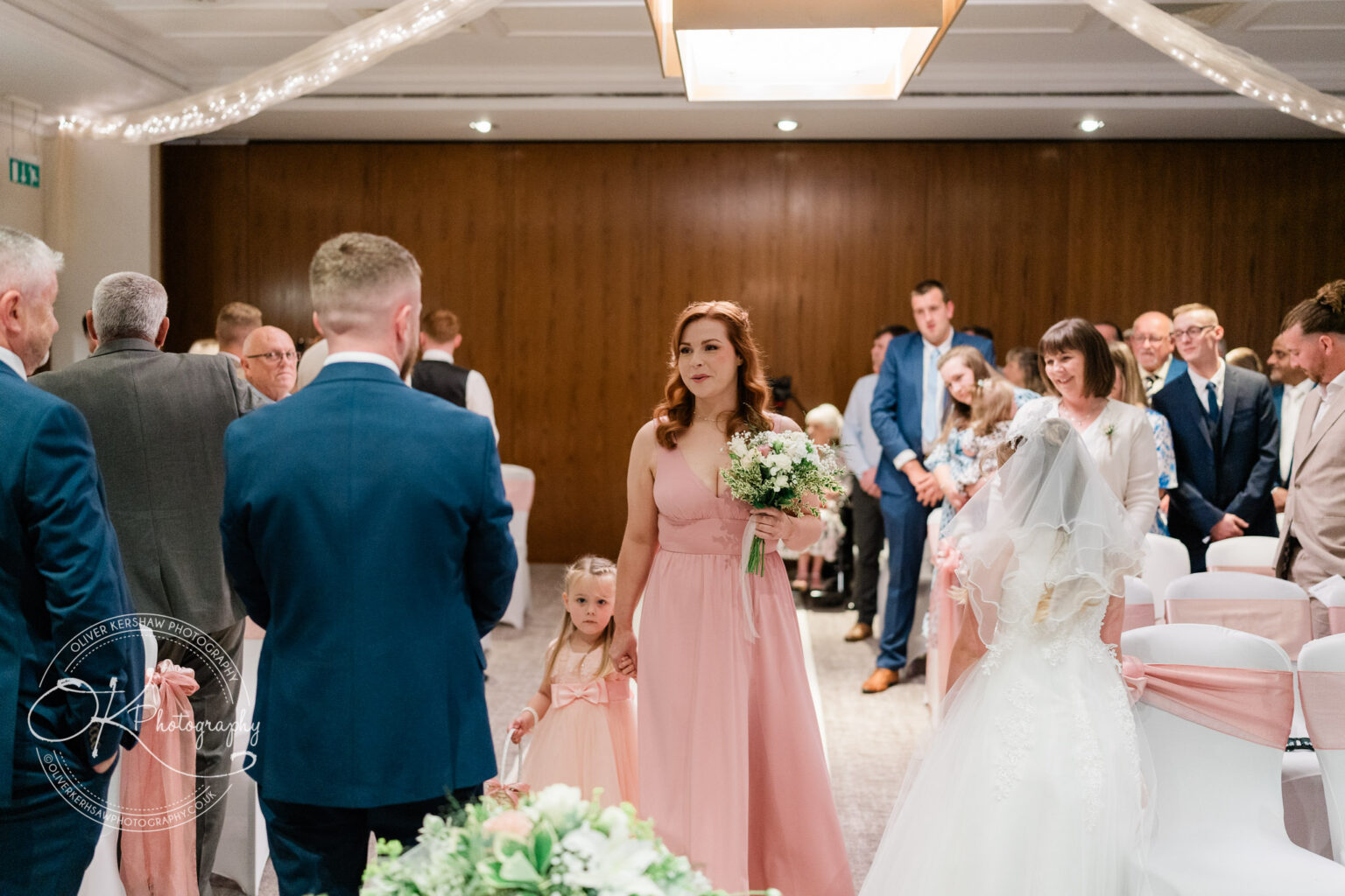 Bridesmaid in a pink dress holds hands with a young flower girl and a bouquet of flowers as they walk down the aisle at a wedding ceremony, with guests seated and standing in the background.
