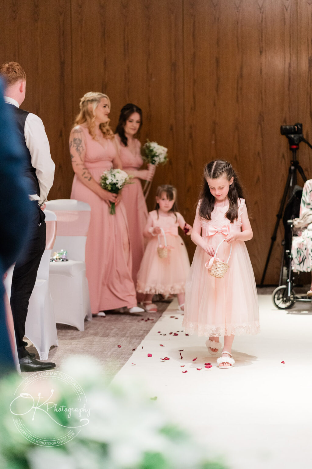 Two young flower girls in pink dresses walk down the aisle scattering petals, followed by two bridesmaids holding bouquets.