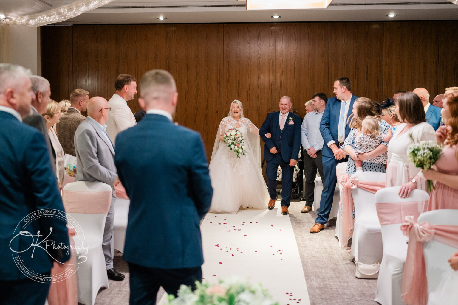A bride in a white wedding dress walking down the aisle with her father, surrounded by seated guests in a decorated venue.