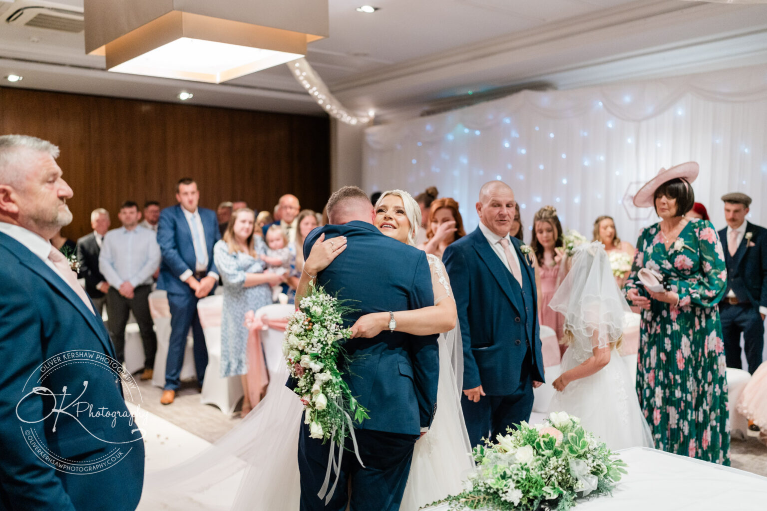 A bride hugging a groom at a wedding ceremony surrounded by guests, with floral arrangements and decorations in the foreground.