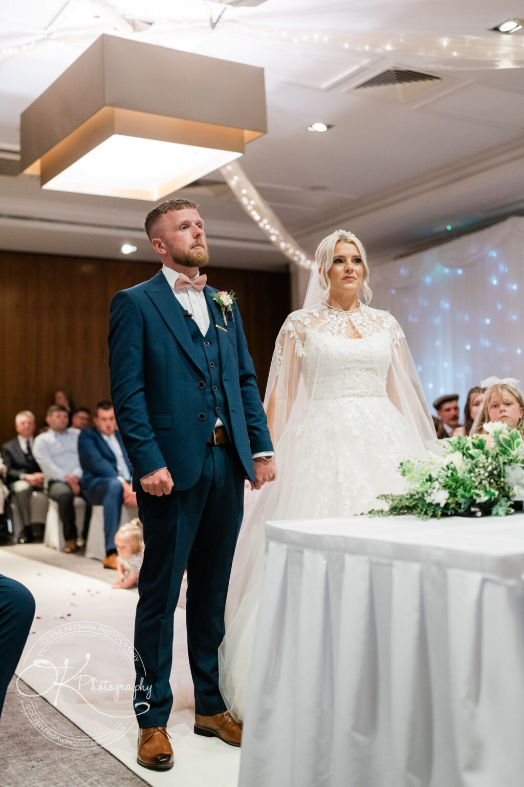 Bride and groom standing together during a wedding ceremony, with the bride in a white lace dress and the groom in a blue suit.