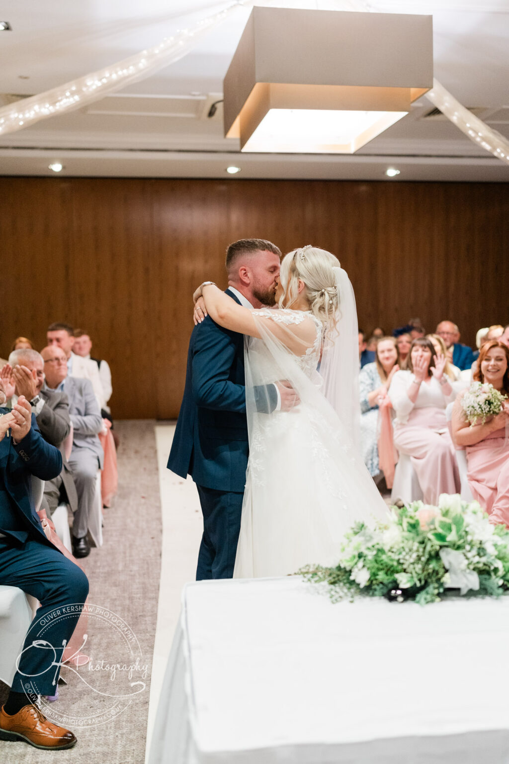 Bride and groom kissing at their wedding ceremony, surrounded by applauding guests.