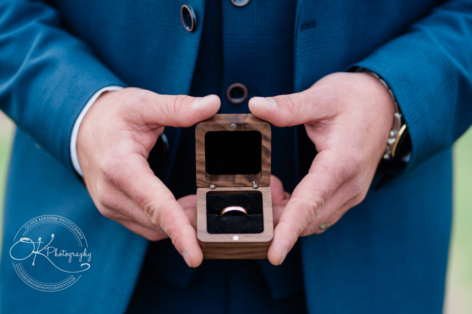 Hands holding an open wooden ring box with a gold wedding ring inside.