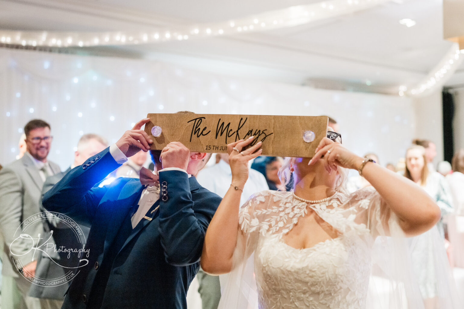 Bride and groom holding a sign reading "The McKays 15th June" at their wedding reception, with guests visible in the background.