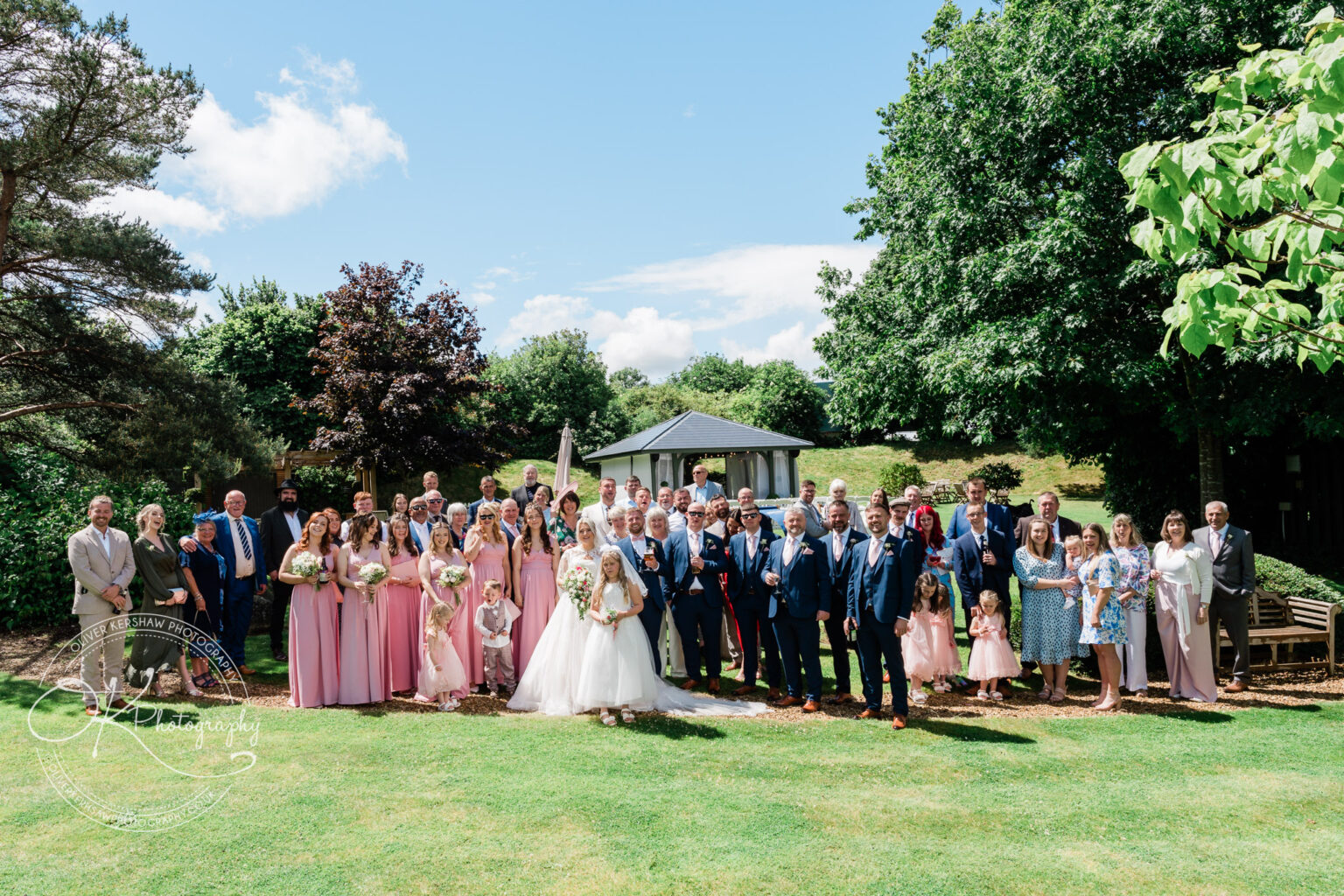 A large wedding party group posing outdoors in a garden setting, with bridesmaids in pink dresses and groomsmen in blue suits.