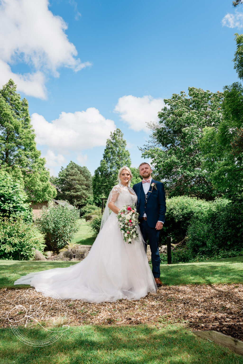 Bride and groom standing together in a garden, with the bride wearing a white wedding dress and the groom in a navy suit, under a blue sky with scattered clouds.