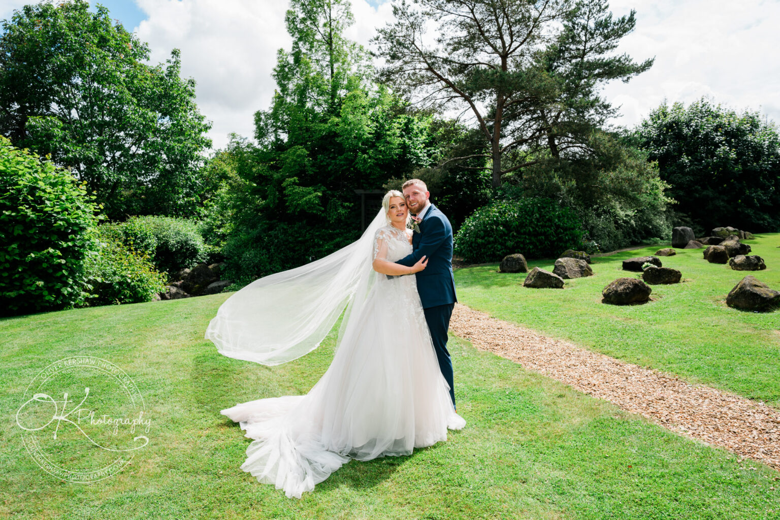 Bride and groom in wedding attire embrace on a lush green lawn with trees and rocks in the background.