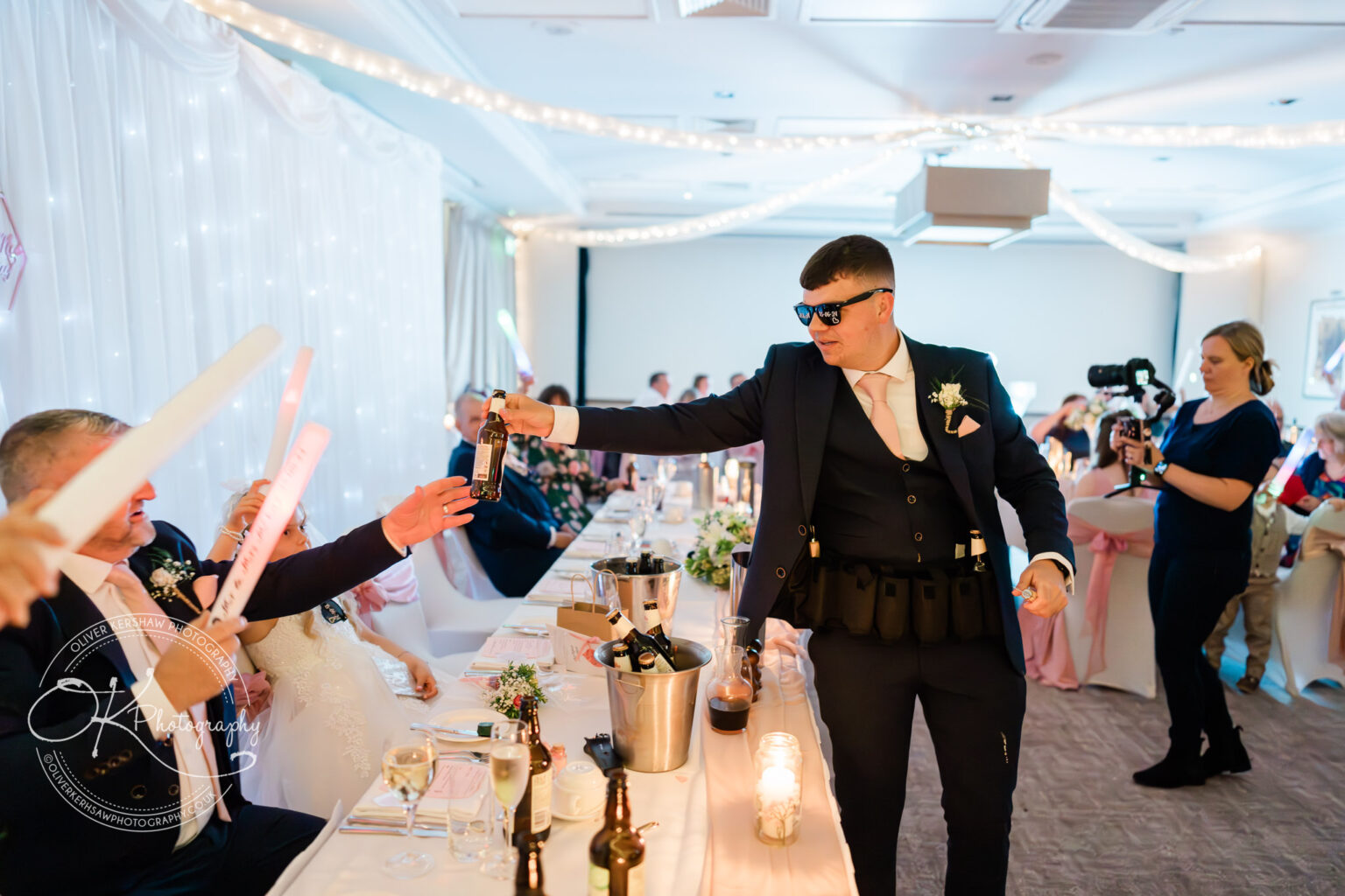 A man in a suit and sunglasses hands a beer to a man at a wedding reception while a photographer captures the moment.
