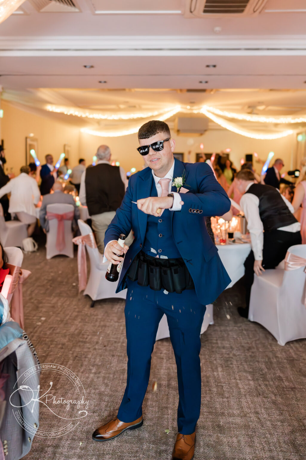A man in a blue suit and sunglasses holding a bottle and partying in a decorated reception hall with guests and colourful lights in the background.