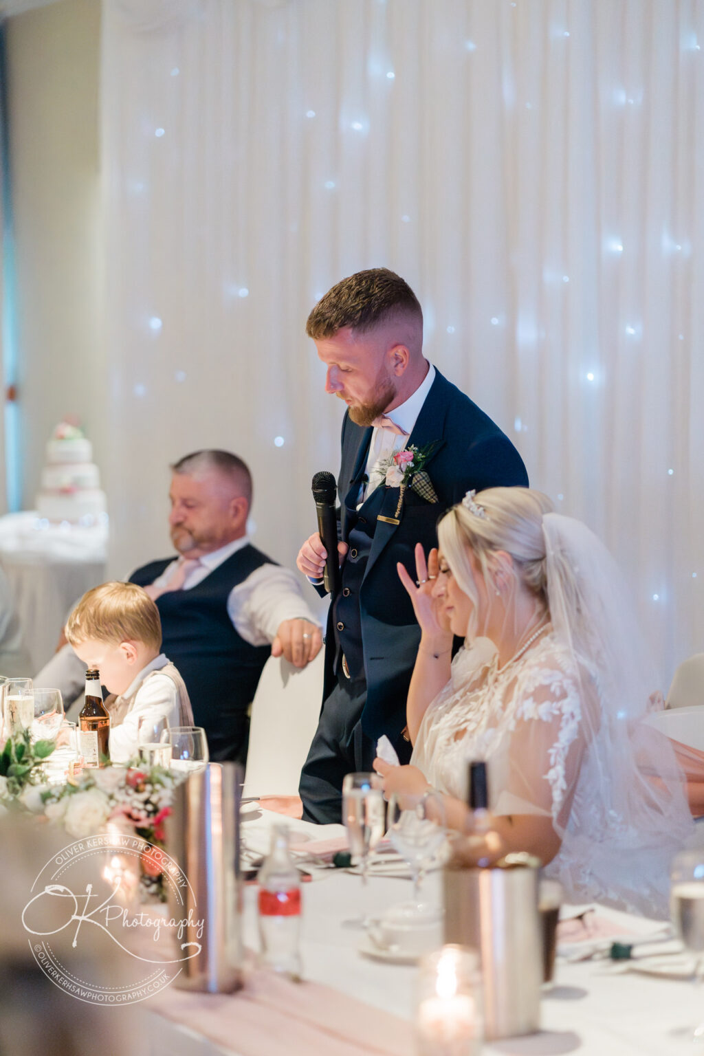 A groom in a navy suit gives a speech at a wedding reception, holding a microphone. He stands next to his bride, who is seated and wiping her face with a napkin. A child and an older man are also seated at the table. There are floral decorations, candles, and drinks on the table, and a wedding cake is visible in the background.