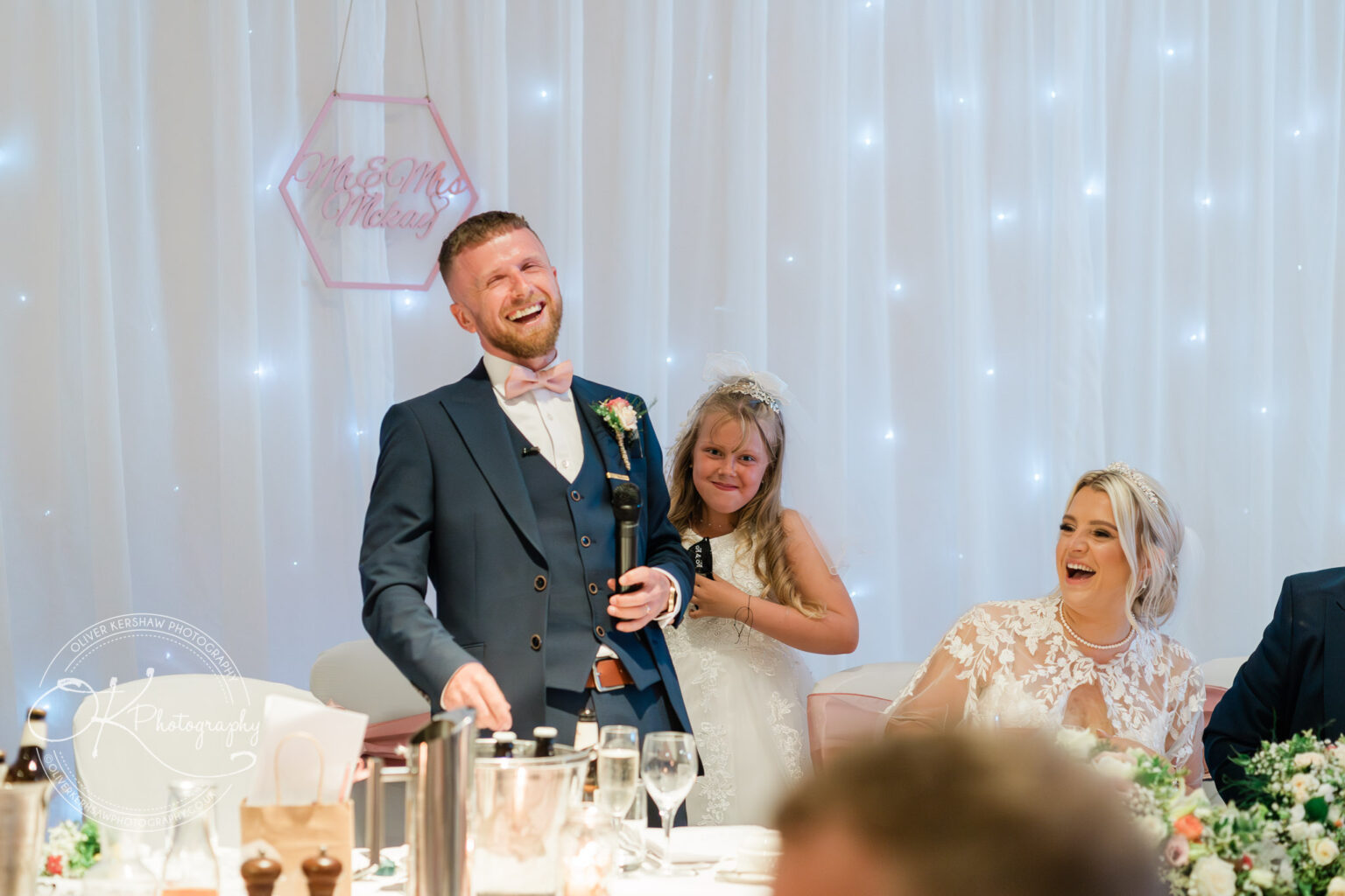 Man in a suit and bow tie laughs while holding a microphone, with a young girl and a woman in a lace dress smiling beside him at a wedding.