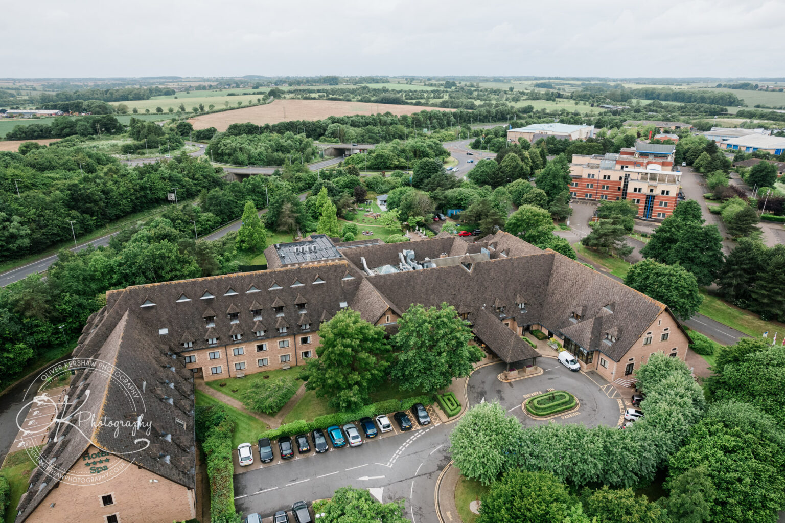Aerial view of a large, thatched-roof building surrounded by trees and parking spaces, with more buildings and open countryside in the background.