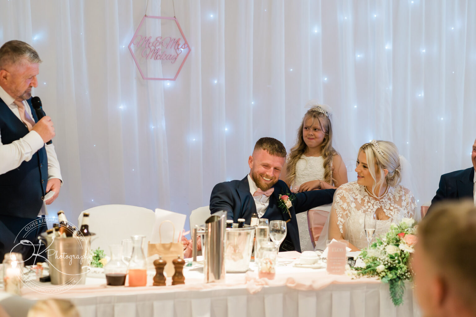 Man giving a wedding speech to a seated newlywed couple with a young girl standing behind them, surrounded by decorations and twinkling lights.