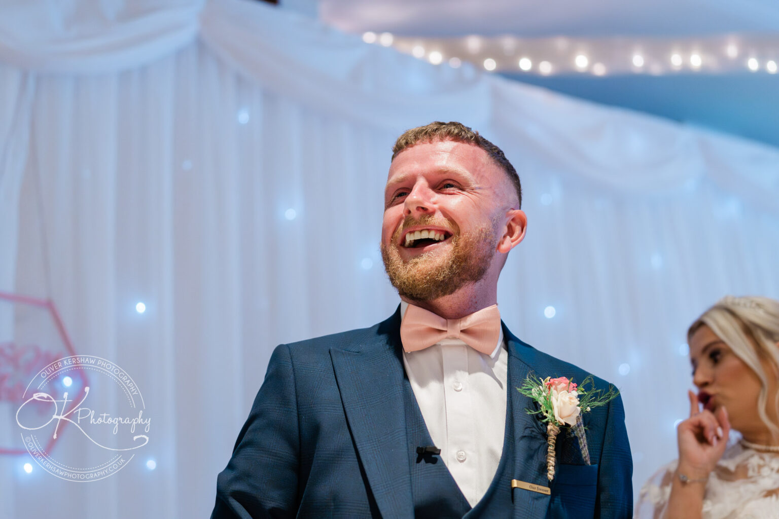 Man in a blue suit with a pink bowtie laughing, with a woman in the background against a backdrop of white drapery and fairy lights.