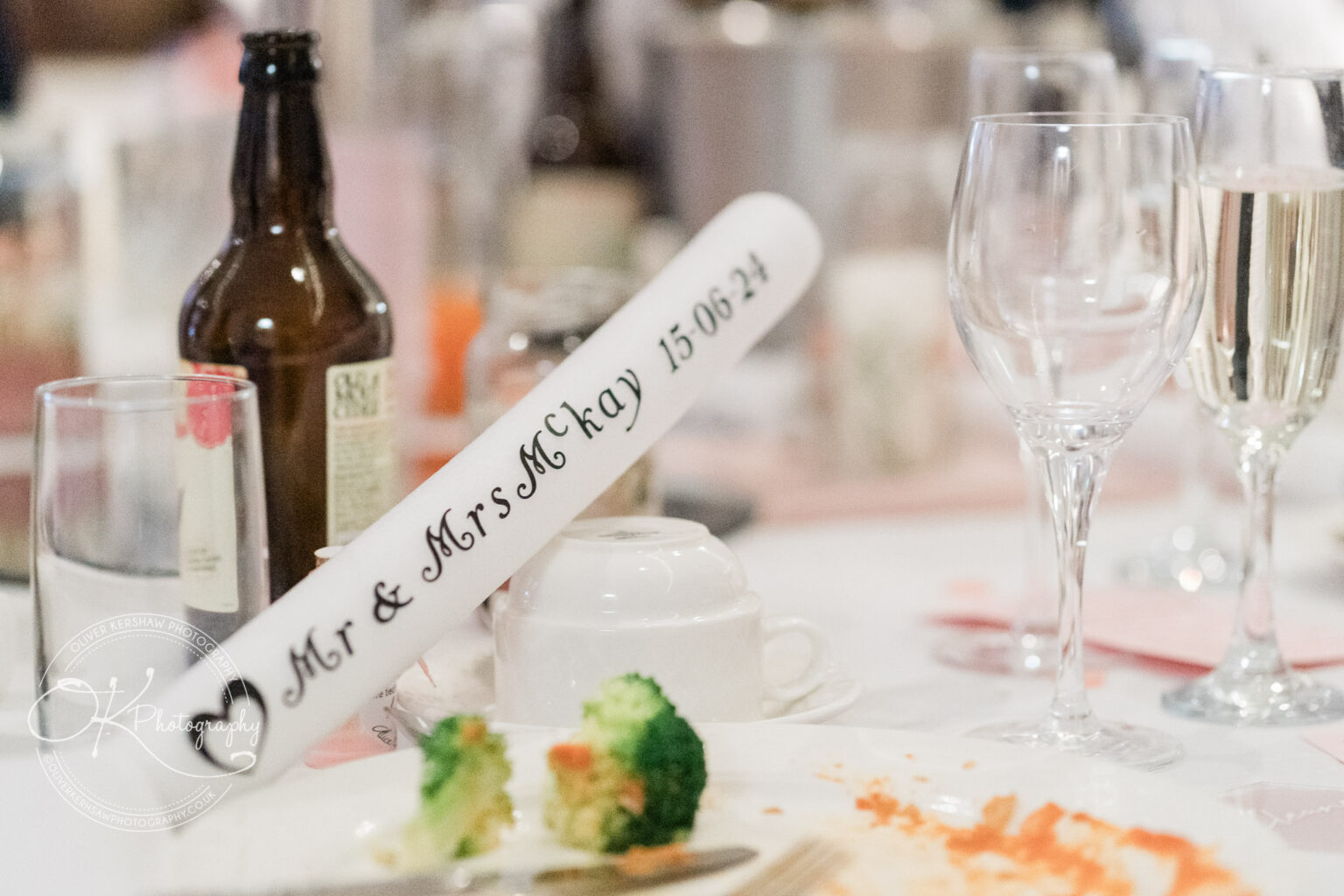 Table setting with wine glasses, a beer bottle, a white teacup, and a scroll reading "Mr & Mrs McKay 15-06-24".