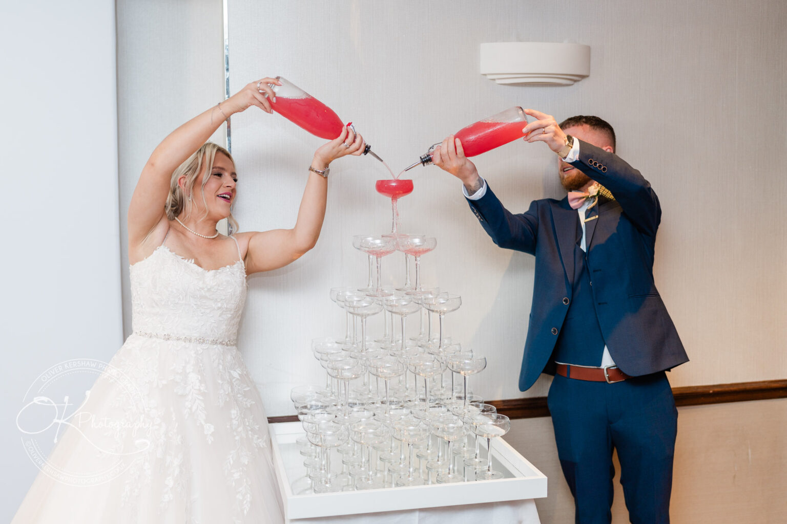 Bride and groom pouring red drinks into a pyramid stack of champagne glasses.