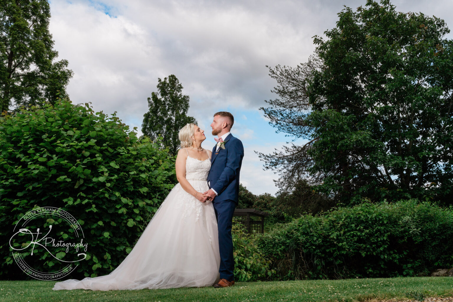 Bride and groom holding hands and gazing at each other outdoors, surrounded by greenery.