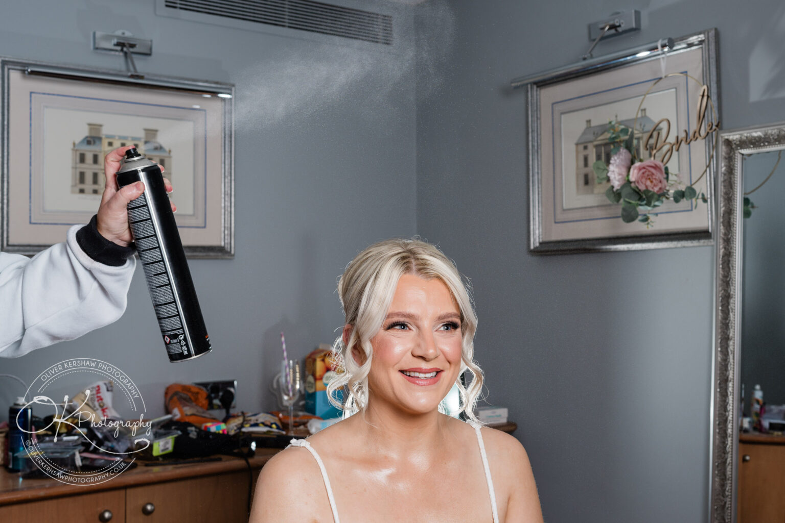 A smiling woman getting her hair sprayed in a dressing room, with the word "Bride" in the background.