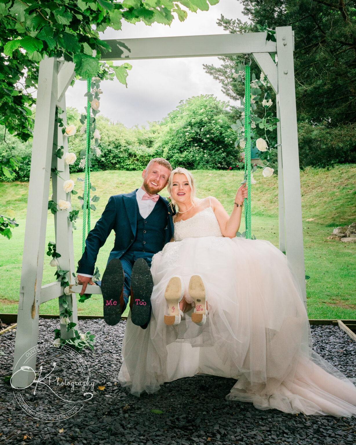 Bride and groom in wedding attire sitting on a swing with "Me Too" and "I Do" written on the soles of their shoes.
