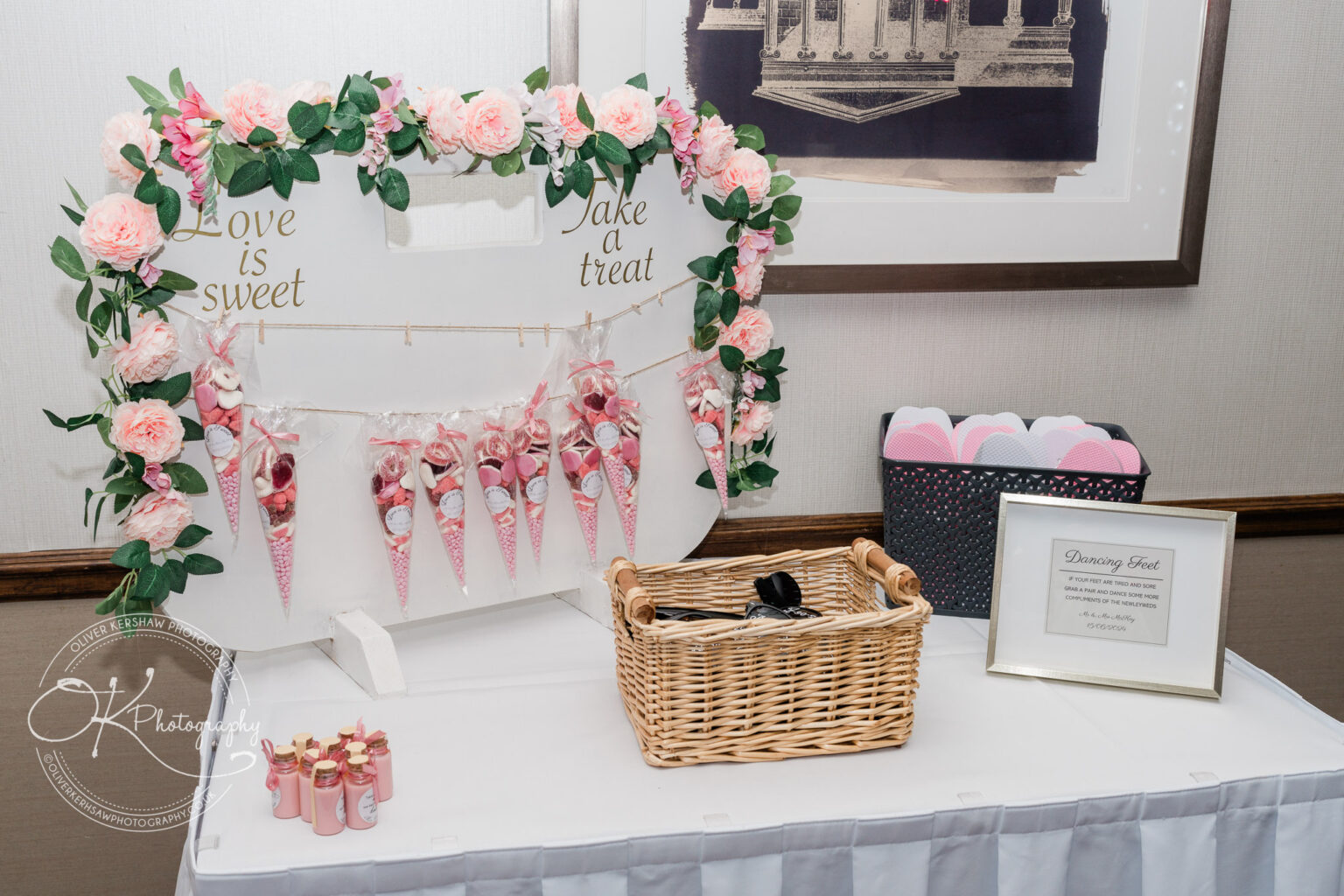 Wedding candy table adorned with pink flowers, featuring a sign "Love is sweet" with candy-filled cones hanging below. Nearby, a basket of flip-flops is accompanied by a framed sign offering them for "Dancing Feet".
