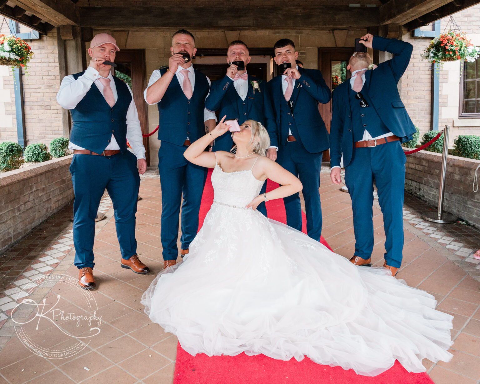 Bride in a white wedding dress kneeling on a red carpet, surrounded by six groomsmen in blue suits, all drinking from flasks.