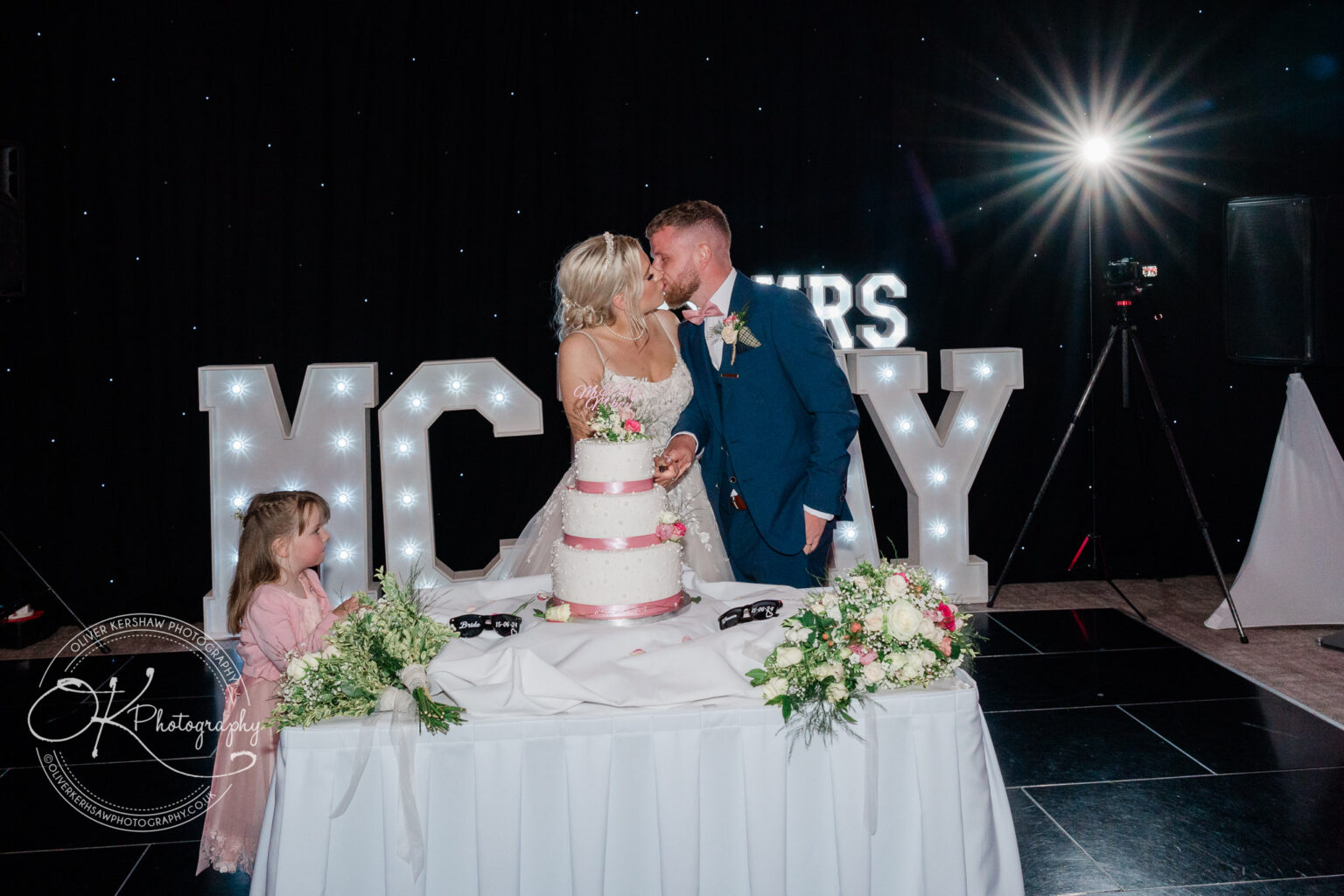 Bride and groom kissing while cutting wedding cake, with a young girl watching, in front of large illuminated letters.