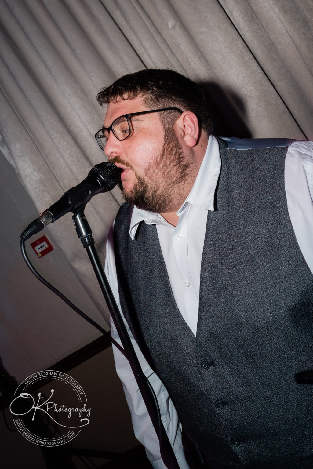 Man wearing glasses, a white shirt, and a grey vest singing into a microphone.