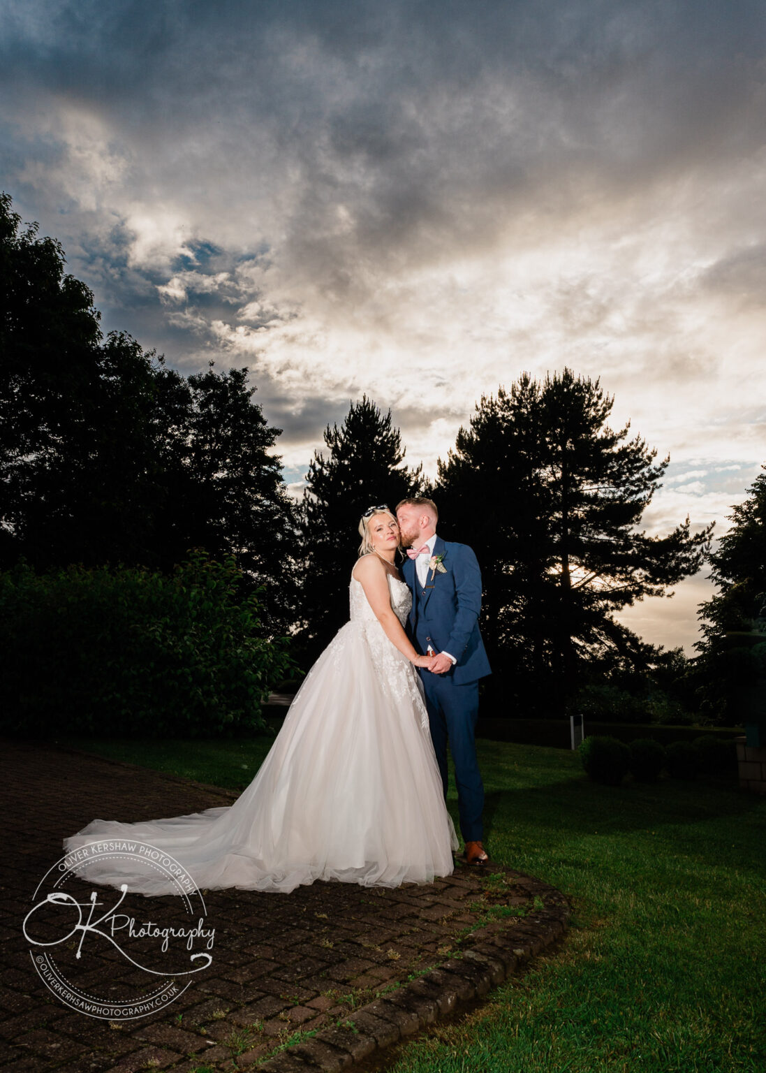 Bride and groom in a garden at sunset, with dark trees and a cloudy sky in the background.