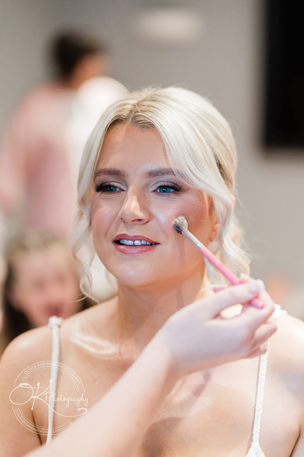 A woman with blonde hair is getting her makeup done, with a focus on applying blush on her cheek with a makeup brush.